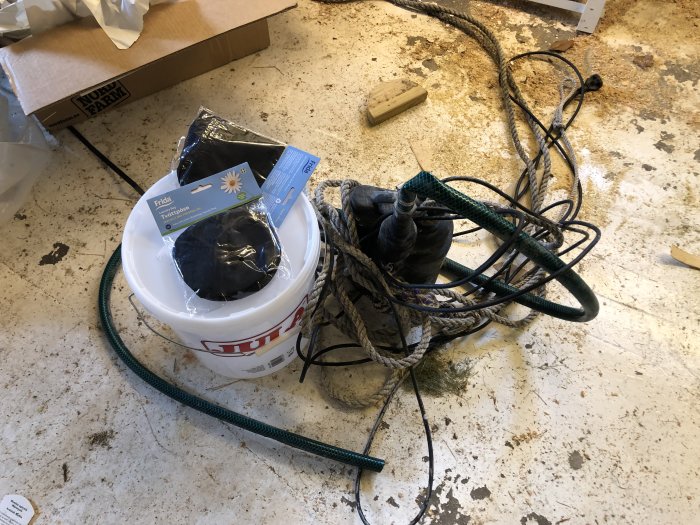 Prototyp av skimmersystem med hink, dränkpump, tvättpåsar och rördelar på en smutsig verkstadsgolv.