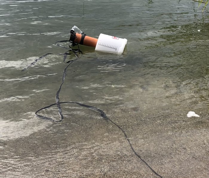 En apparat med rör och flytelement delvis flytande i vatten vid strandkant.