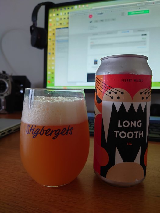 Ett glas fyllt med grumlig öl bredvid en färgglad ölburk med texten "LONG TOOTH IPA", framför en datorskärm.