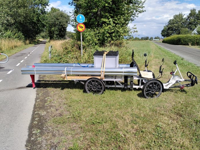 Cykel med lång släpvagn lastad med takavvattningssystem vid vägkant.