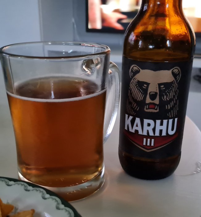 Ölglas fyllt med bärnstensfärgad dryck bredvid en flaska med björnlogotyp, i bakgrunden en oskarp TV-skärm.