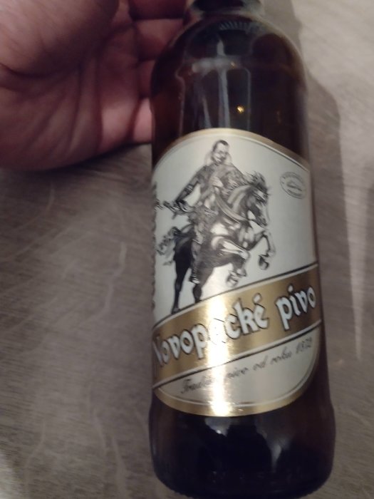 Hand håller en flaska Tjeckisk öl med etikett som visar ridande krigare, "Novopacké pivo". Bilden är suddig.
