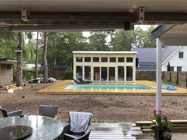 Utsikt från en terrass mot en pågående husbyggnation med ett halvfärdigt vitt hus och en nylagd träterrass.