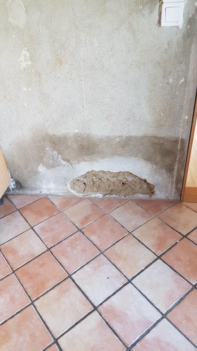 Damaged plaster on an interior wall, showing a patch of exposed brick near the floor. The wall has uneven texture and discoloration.