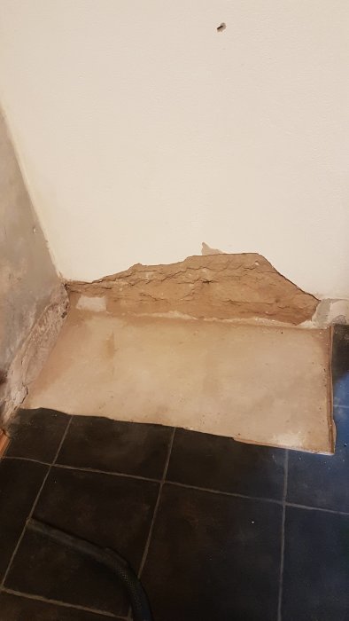 A damaged wall corner in a house with missing plaster, showing exposed brown and beige layers, near a black tiled floor.