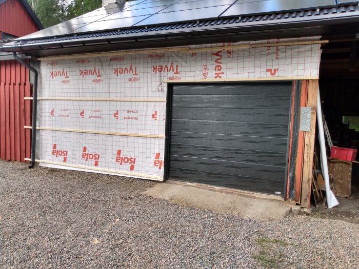 Vägg av garage under renovering med ny vindskyddsisolering och en stängd svart garageport.