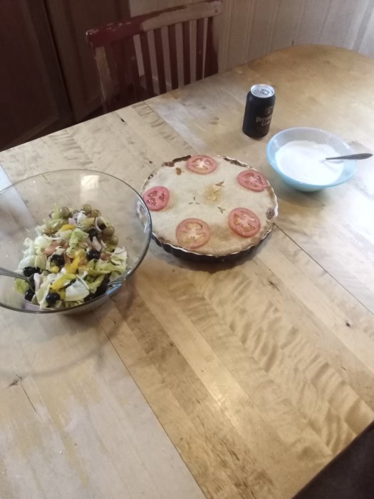 Tacopaj på bord med skivade tomater ovanpå, sallad med oliver och rödlök bredvid, vitlökssås och en läskburk.