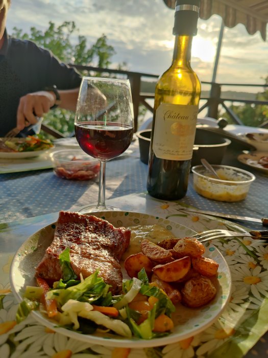 Middag utomhus med grillat kött, rostade potatisar, sallad, ett glas rött vin och en vinflaska i solnedgång.