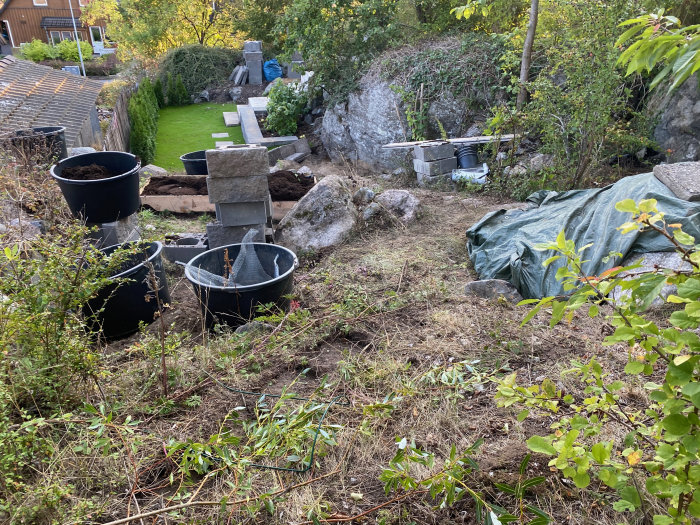 En trädgård under anläggning med grävda områden, hinkar fulla med jord, sil och en presenning som täcker marken.