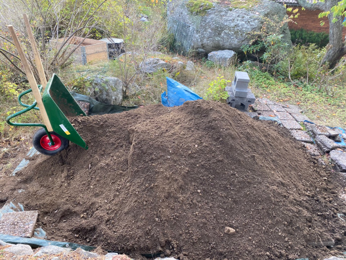 Stor jordhög bredvid skottkärra och stenplattor med presenning och mur i bakgrunden på en trädgård under förberedelse.