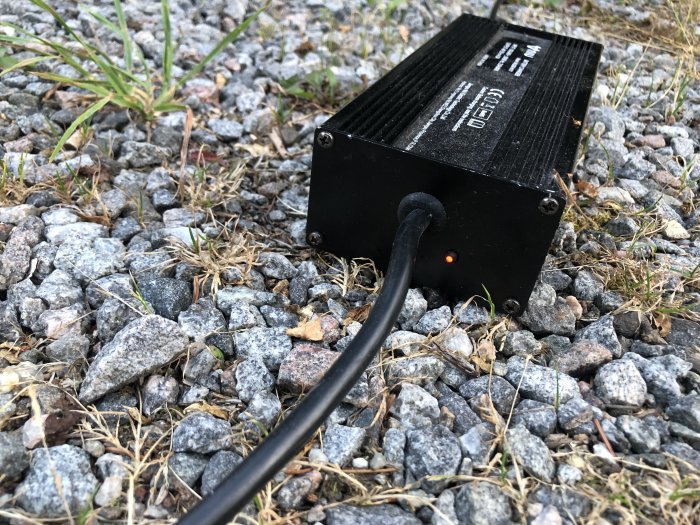 Svart BMS (Battery Management System) med röd lampa som lyser på grusunderlag.