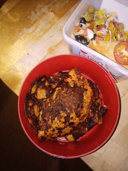 Stekta pannkakor i en röd skål och en matlåda med sallad på ett slitet träbord.