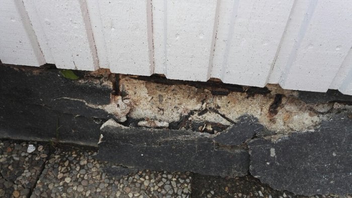 Skadad grund på 70-talshus med lossnande bitar och flagnande svart färg vid husvägg.