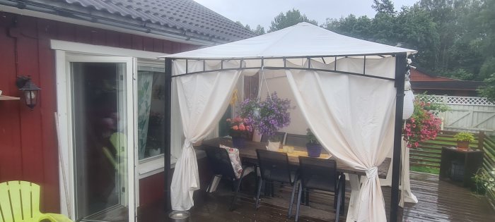 En befintlig altan med ett vitt, temporärt tälttak, närliggande hus och garage, blommor och trädgårdsmöbler.