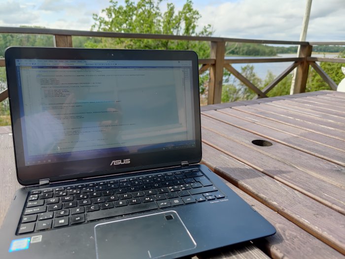 Laptop öppen på kodningssida på ett uteplatsbord med utsikt över en sjö i lantlig miljö.