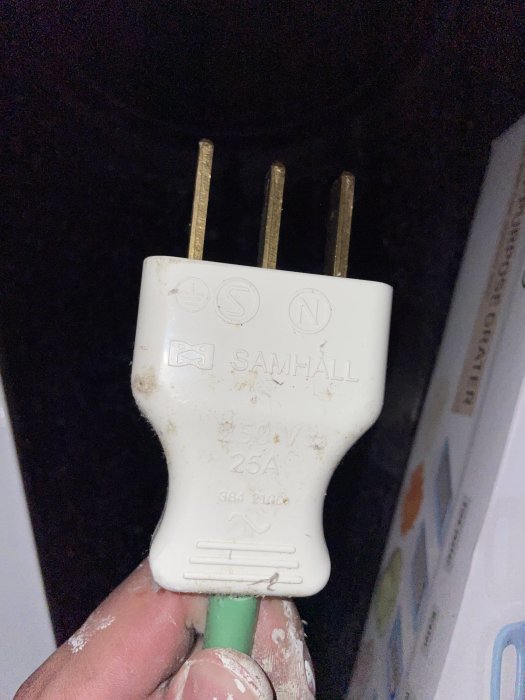 Hand håller en smutsig vit elkontakt med texten "SAMHALL 250V 25A" och elsymboler, mot en svart bakgrund.