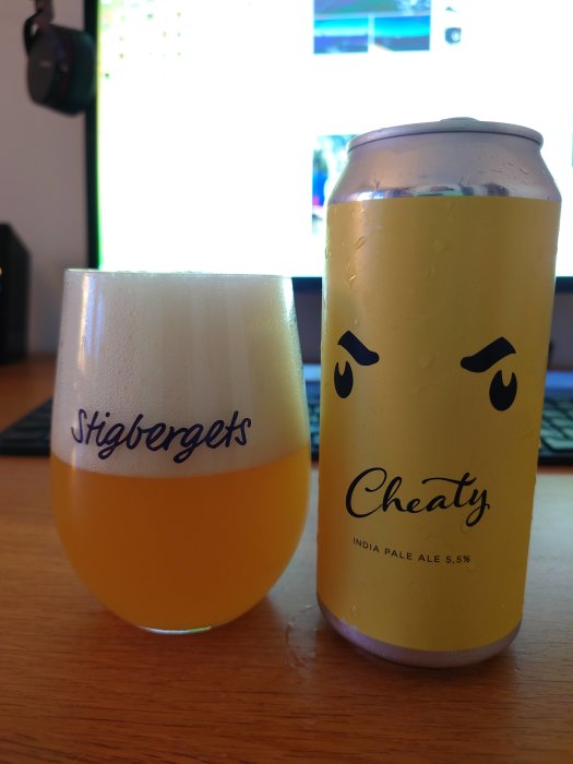 Ölglas fyllt med hazy DIPA bredvid en burk IPA medans en skärm syns i bakgrunden.