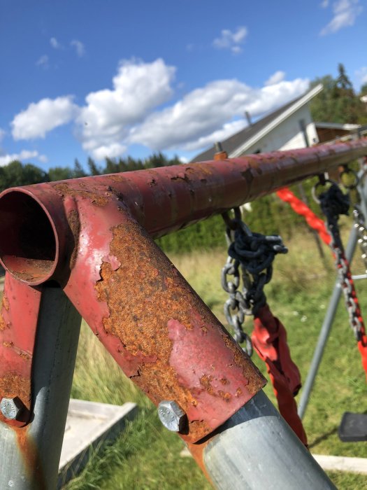 Rostig del av en gammal gungställning i förgrunden med grönska och hus i bakgrunden.