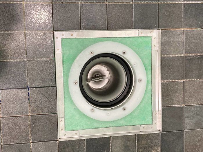 Öppen golvbrunn med Tile in slim insats, omringad av mörkgrå kakelplattor och grönt tätskikt.