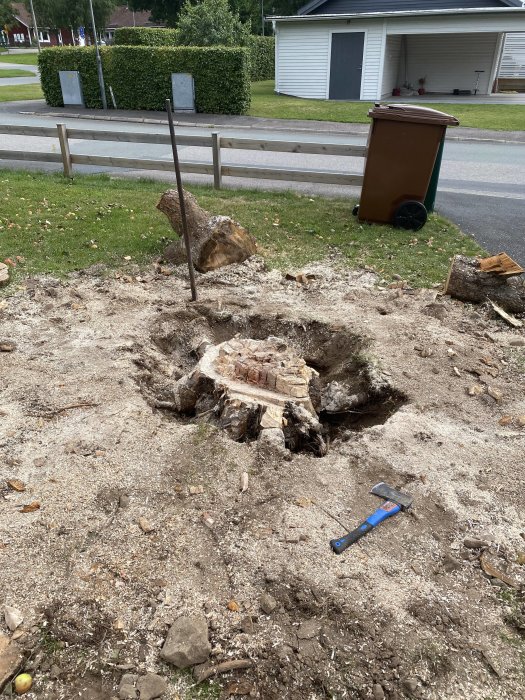 En stor trädstubbe med rötter och en yxa på marken vid ett bostadsområde.