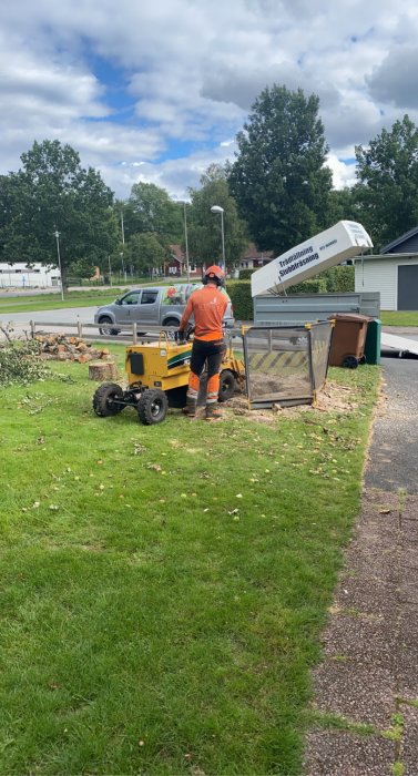 Arbetsperson i orange klädsel använder stubbfräs på en trädgårdstomt.