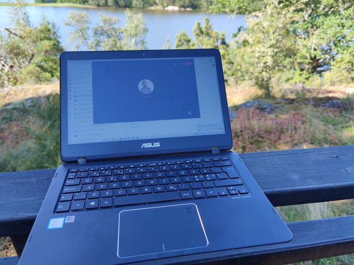 Laptop på ett höj- och sänkbart bord med utsikt över en sjö och grönska i bakgrunden.