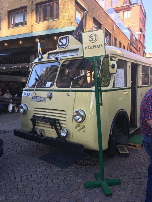 Gammal veteranbuss märkt "KLUBB GAMLA ÖSTEN" parkerad vid en torghallplats, med svensk flagga och skylt.