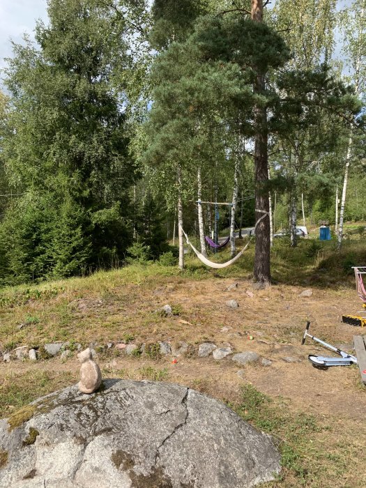 Ojämn skogstomt med stenar och buskar, en hängmatta mellan träd och leksaker på marken.