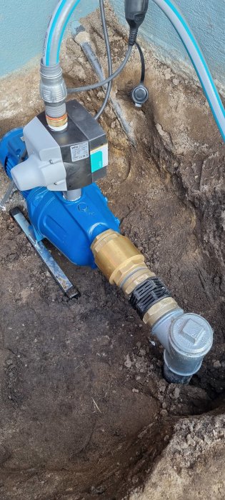 En nyinstallerad blå vattenpump kopplad till rörsystem i en grävd grop, med synliga kablar och slangklämmor.