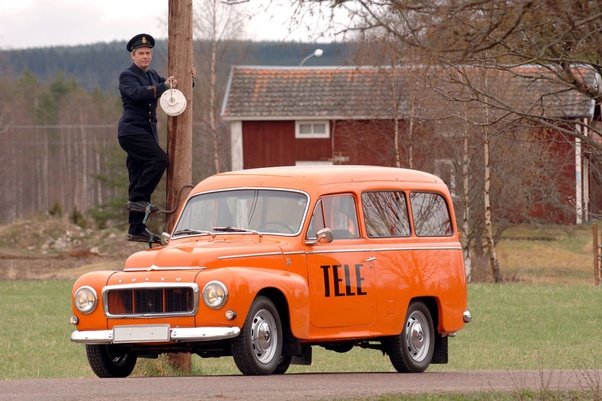 En person i Televerkets uniform klättrar på en telefonstolpe vid en orange Tele-servicebil.