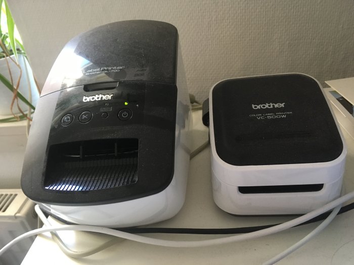 Två etikettskrivare från Brother, en svart och en vit, på ett bord redo för användning.