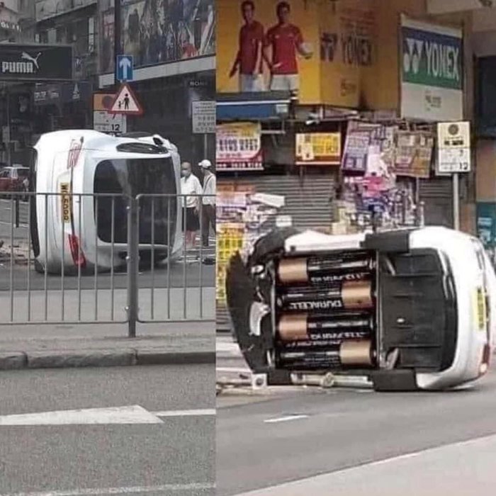 Vit minivan ligger på sidan liknande en omkullvält batteri, på en stadsgata med bakgrund av affärer.