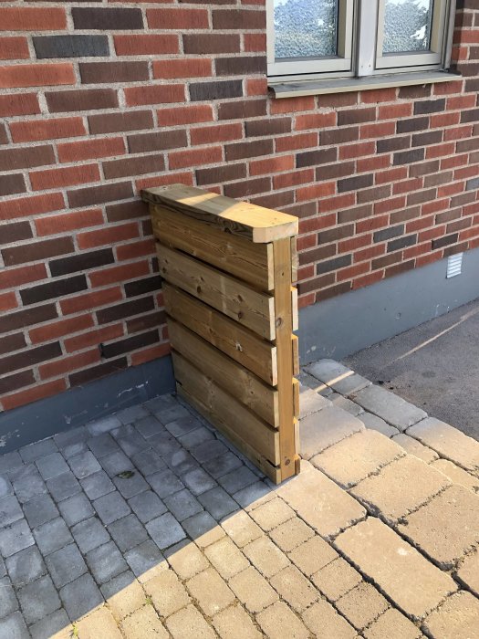 Pystysuora puuaita tiiliseinän edessä, ruskeasta painekyllästetystä puusta, seisoo laatoitetulla pihalla.