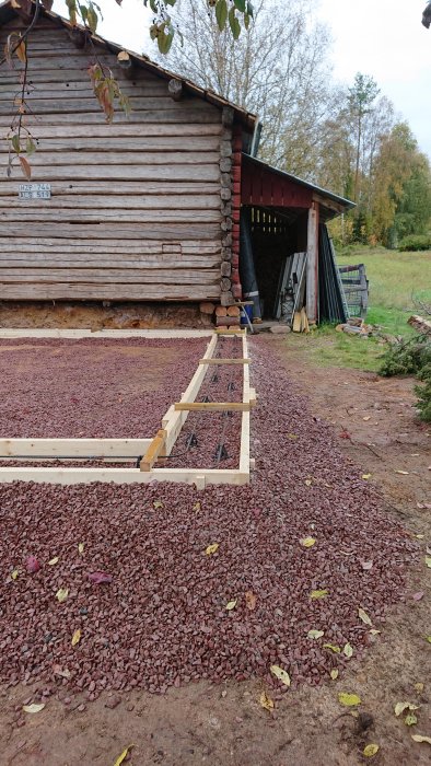 Nygrundad betongplatta för timmerstomme bredvid carport, med makadamunderlag och träramar.