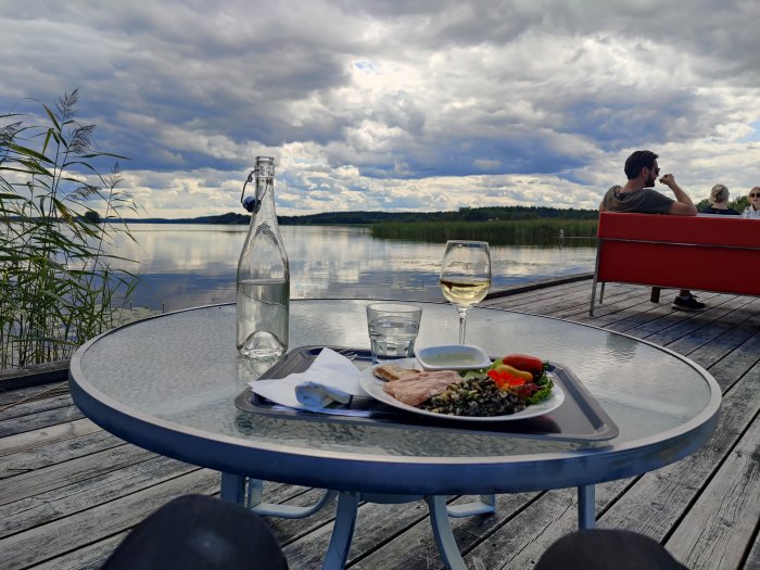 Lunch på ett bord vid en sjö med en vinflaska och ett glas, med personer som samtalar i bakgrunden.