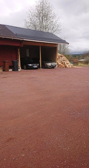 Hög carport med tak av tp20-plåt, snörasskydd, och timmerpanel på baksidan, intill en vedhög.