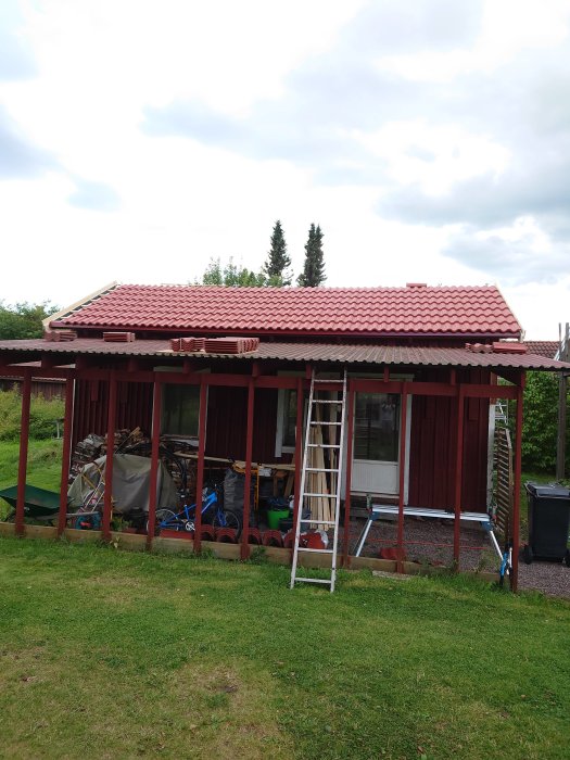 Ett nästan färdigställt rött tegeltak på ett uthus med stege lutad mot taket och byggmaterial synligt.