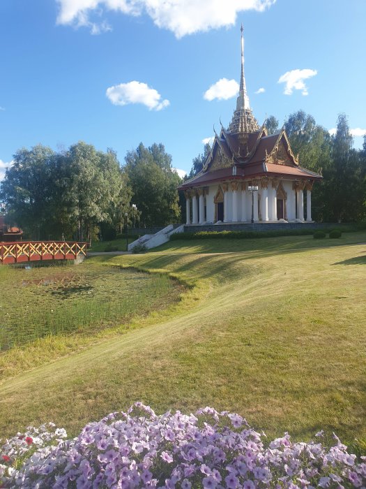 Thailändskt tempel i exotisk stil med spetsiga tak och gyllene detaljer omgivet av grönska och blommor.