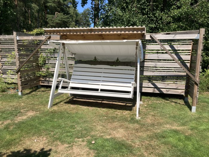 Vit hammock med takställning vid ett träplank, placerad på en solig gräsmatta med skog i bakgrunden.