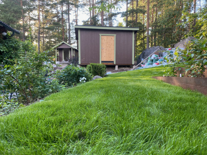 Nybyggd redskapsbod med oljad dörr, grön gräsmatta i förgrunden, och utegrävd jord runtom.