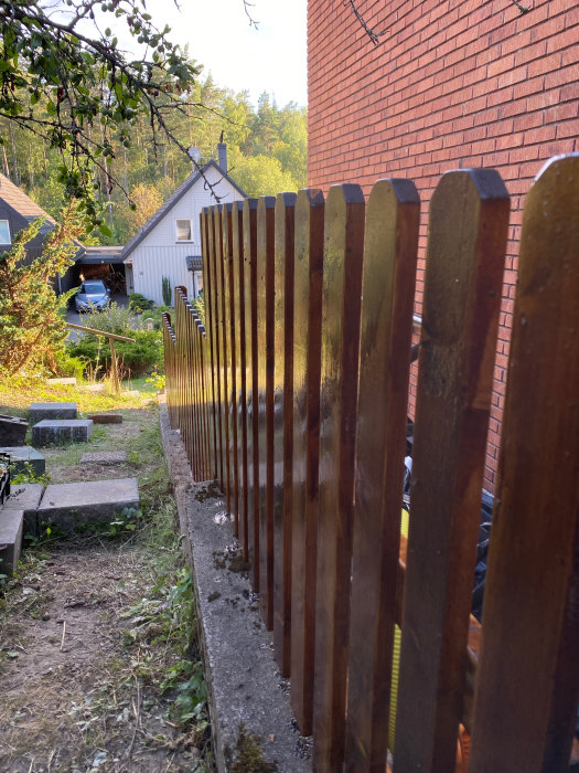 Nymålat trästaket i brun tjärlik färg vid en trädgård med utsikt över hus och skog.