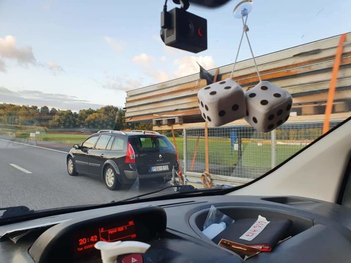 Bil med öppen baklucka och virke som sticks ut, sett från ett annat fordon med tärningar hängandes.