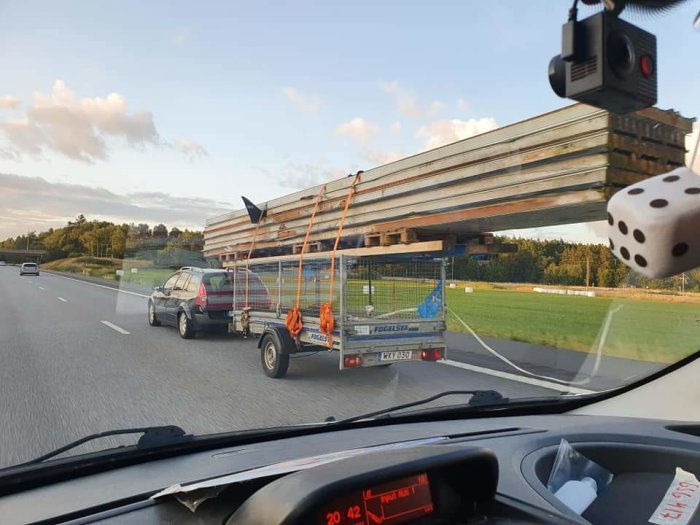Bil med släp överlastat med långa metallbjälkar på motorväg, potentiellt farlig lastning.
