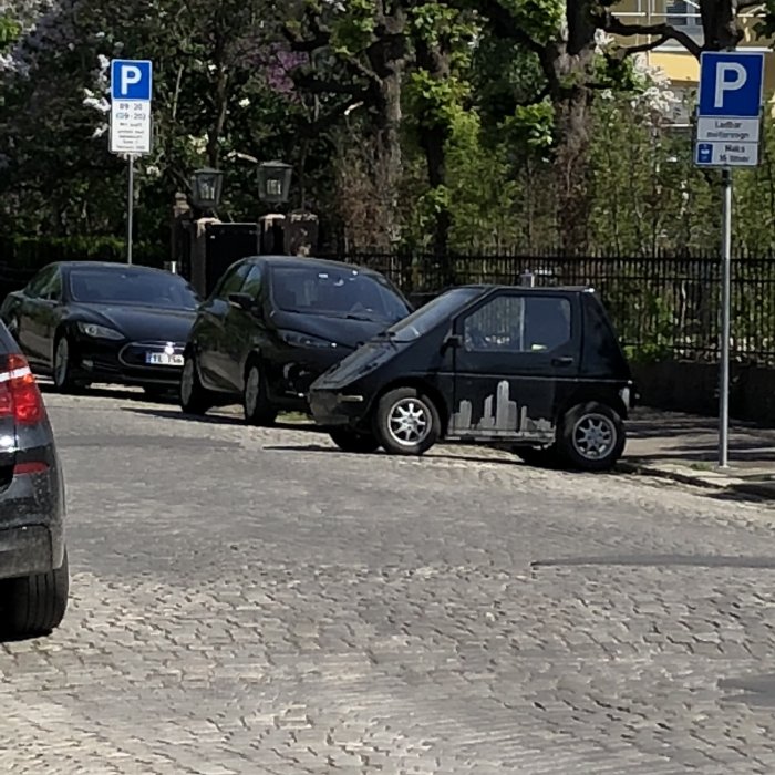 Bilar parkerade på en gata med kullersten, en mycket liten bil framför.