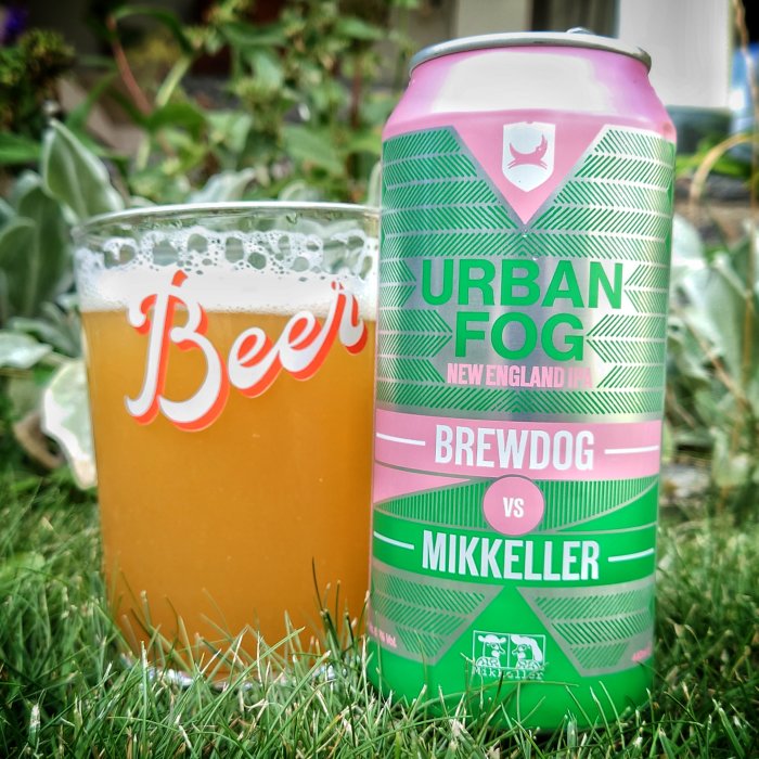 Burk av Urban Fog New England IPA av BrewDog vs Mikkeller bredvid ett glas öl på gräsmatta.