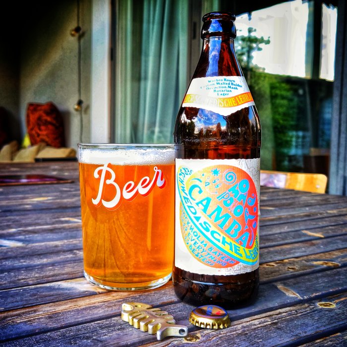 En öppnad ölflaska och ett glas med öl på ett träbord, med en kapsylöppnare och kapsyl.