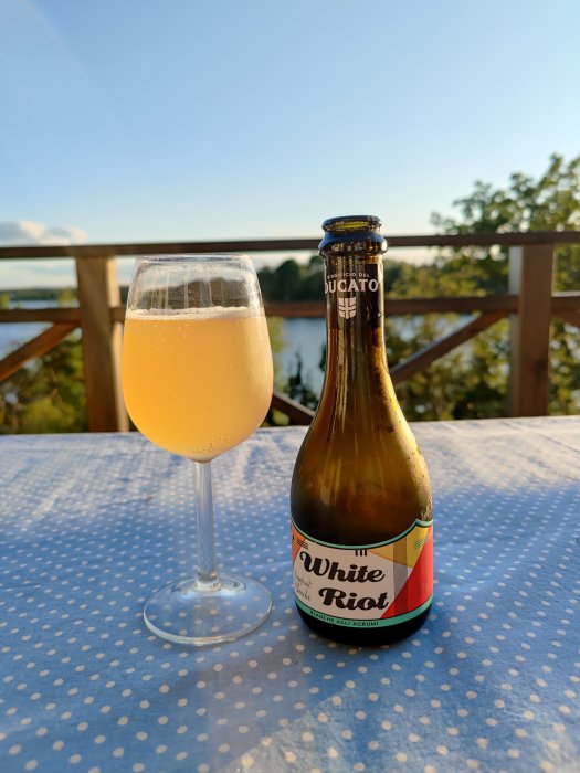 Flaska av "White Riot" veteöl bredvid ett glas fyllt med öl på ett bord med blåvit duk, med natur i bakgrunden.