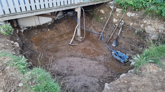 Grävd grop på 1,6 meters djup för byggprojekt med verktyg och vattenansamling, nära byggnadsgrund.