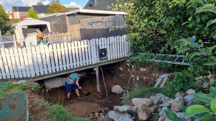 En person gräver en stor grop nära en staket vid ett hus, med högar av jord och sten.