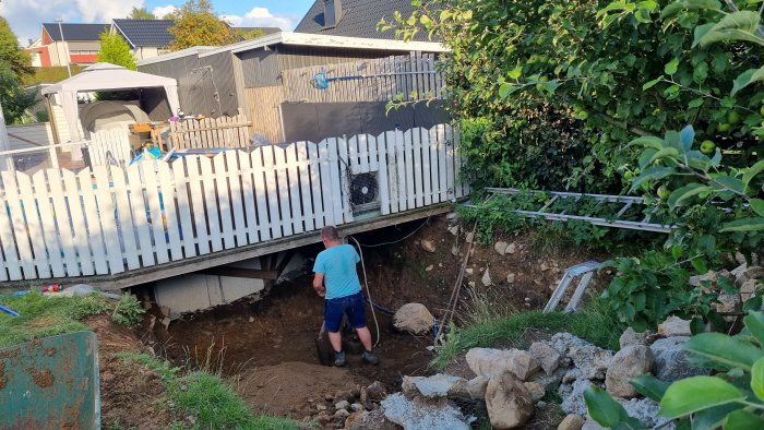 Person gräver stor grop nära husgrund för gjutning av betongplatta, omgiven av sten och utrustning.